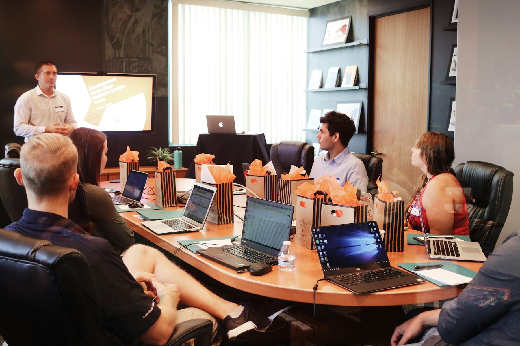 A real meeting: someone presents at a screen while colleagues sit around the table with laptops.